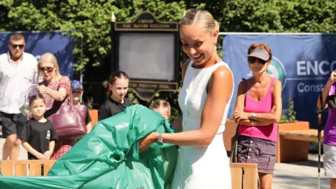 Cowshed PR Boyesen stood next to Dowden, who is wearing a white dress with her blonde hair tied back. They are pulling back a green cover to unveil the intricate 3D map featuring figurines and various places in Caerphilly. There are people clapping in the background watching on.