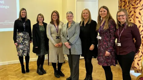 City of Wolverhampton Council Seven women are smiling whilst they are stood posing for a photograph. Behind them is a projector screen, displaying a presentation.