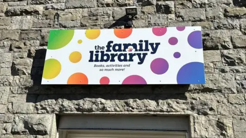 A stone wall with a sign which reads "The Family Library: books, activities and so much more!" It is white with multicoloured spots.