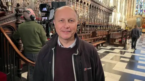 Qays Najm/BBC Peter Taylor standing in King's College Chapel. He is looking directly at the camera and smiling. He is balding and is wearing a black fleece over a dark jumper and checked shirt. Behind him are the choir stalls and the black and white tiled floor of the chapel.