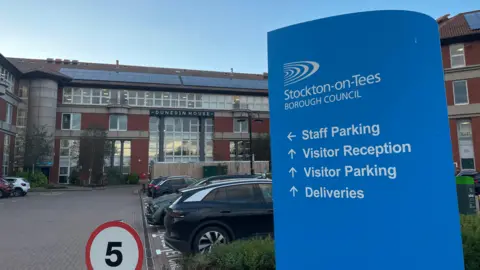 LDRS Brick and glass building with a blue and white Stockton-on-tees Borough Council sign. There are parked cars and block paving.