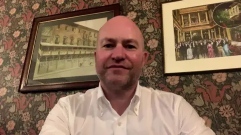 A man with a shaved head and neat stubble, who is wearing a white button-down shirt, sits in front of a wall decorated with dark, floral wallpaper and two framed paintings, one of what looks like a 19th Century building, and the other of an early 20th Century ball.