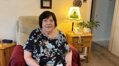 Rita is sitting in an armchair holding a piece of paper in her hand. She is wearing a black top with blue flowers. Behind her is a table with a telephone, lamp and plant. 