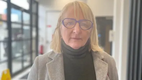 A woman is looking into the camera. She wear purple glasses and is wearing a grey jumper and coat. She has bobbed blond hair. The background is blurred and she is in focus.