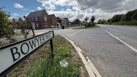 Glenn Pringle/BBC A street view of the A196 which is a single carriageway concrete road. There is a street sign in the bottom left on a grassy verge which reads 'Bowyer Way'. To the left of the road there are new-build red brick houses with another road running in front of them, adjacent to the A196. The sky is blue with large fluffy clouds.