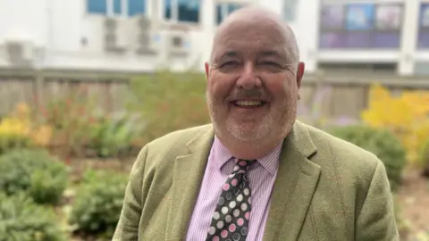 Mark Goldsack smiling into camera in front of shrubbery. He has a white beard and is wearing a light green suit jacket, pink and white shirt and multi-coloured tie.