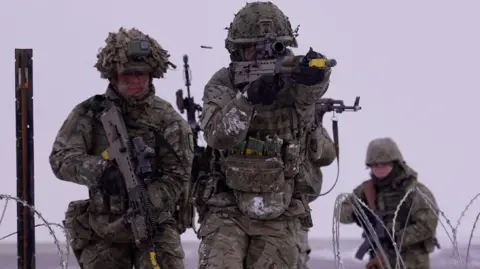 Three soldiers in green camouflage uniforms, holding guns, walking towards a wired fence