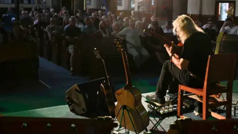 Gordon Giltrap is playing a guitar under a spotlight with other guitars on stands next to him. A packed audience can be seen in the dark beyond the stage sat on church pews.