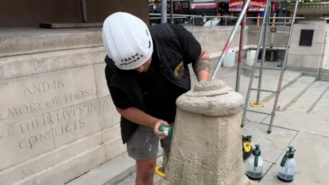 The photo shows a man scrubbing a stone structure. He's wearing a black gilet and a white hard hat. He's holding a green scrubbing brush. There is a stone memorial behind him, and bottles on the floor. 