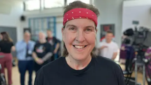 A woman with shoulder length brown hair is smiling at the camera. She is wearing a red bandana with white spots on it, and a black t-shirt