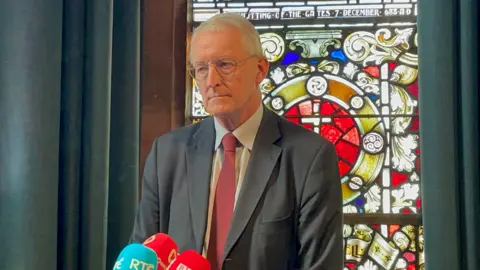 PA Hilary Benn wearing a dark suit and red tei and standing in front of stained glass window at the Guildhall 