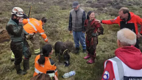 Scarborough and Ryedale Mountain Rescue Team A group of people in high-visability outfits stands around a dog, in an area of moorland. They hold ropes and other rescue equipment.