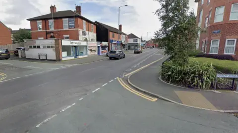 A street view image of a town centre junction. Cars can be seen driving along the road, with shops and business premises on the left. A block of flats is behind a black fence on the right, with a tree near the fence. A street sigh next to the fence says "Marsh Avenue".
