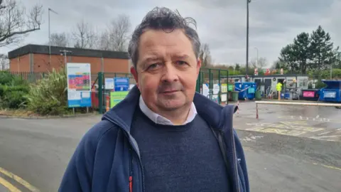 Keith Patterson from the charity Wrap (Waste and Resources Action Programme) at Holywood Recycling Centre in County Down. He has dark grey hair and a blue fleece over a blue jumper and white shirt