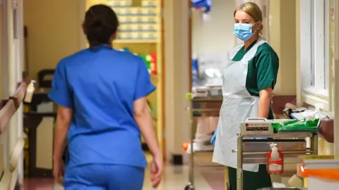 BBC Two women in a hallway in a medical setting wearing face masks and blue scrubs.