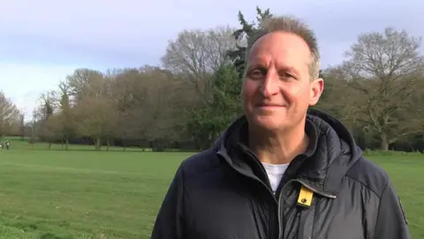 Michael Young smiling at the camera has grey hair and is wearing a black jacket and white t-shirt underneath. Greenery from the golf course in the background.