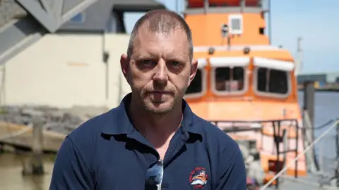 Shaun Whitmore/BBC A man wearing a navy polo shirt and he has sunglasses attached to his shirt. He is looking directly at the camera with the pier behind him 