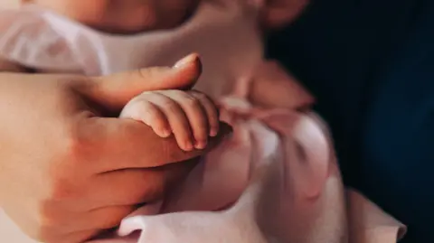 Getty Images A close-up of a mother gently holding her baby's tiny hand, symbolizing love, care, and the precious bond of family. The soft tones and delicate details create a warm and emotional image, ideal for themes related to motherhood, parenting, and family values.