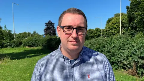 Sam Read/BBC Tom Sisley looks directly at the camera on a sunny day. He has cropped brown hair, wears black rimmed glasses and is wearing a blue and white checked shirt. Behind him is grass and green bushes. 