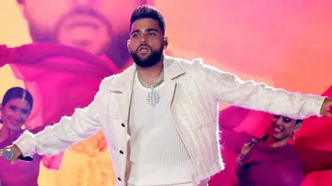 Getty Images A man wearing an all white outfit with his hands out wide as he performs on stage. Behind him are two female dancers wearing pink and purple.