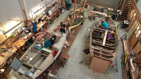 Martin Giles/BBC A bird's eye view of a boatyard workshop. There are three boats being worked on by apprentices. They are all in various states of repair.