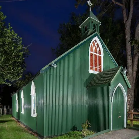 Historic England A green church made from tin with tall, pointed windows and doors surrounded by grass and trees