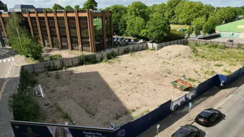 Steve Hubbard/BBC A flattened building site surrounded by hoardings advertising a new housing development.