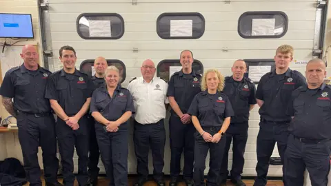 Cambridgeshire Fire and Rescue Service Mr Gooderham is stood in front of the doors used by the fire trucks when they are deployed. With him are other crew members, who are dressed in a blue uniform. 