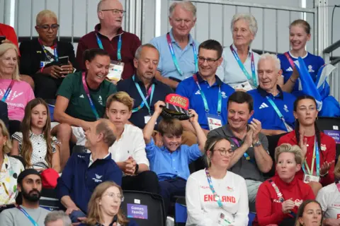 PA Media A crowd at the Sir Chris Hoy Velodrome , including the three royal children Charlotte, George and Louis. Louis is laughing and holding a hat above his head