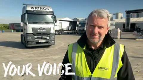 A man stood in a yard with a large white lorry behind him. He is wearing a reflective yellow jacket. He has short grey hair and a short beard.