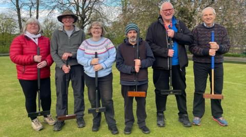 Four men and two women stand smiling and holding up croquet mallets