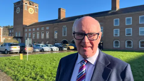 James McInnes, the Conservative leader of Devon County Council, wearing a blue shirt and striped tie with a navy blue blazer, standing in front of County Hall