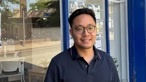 Ray Yu, a young man with short, dark hair, glasses and wearing a blue polo shirt, standing outside a shop.