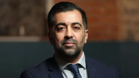 A dark haired man with a dark beard dressed in a dark blue suit with matching tie