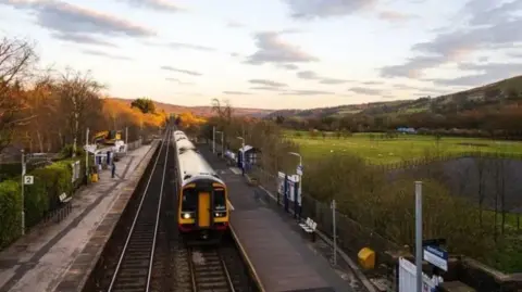 East Midlands Rail A train in a countryside setting with rolling green hills and a dusky sunset. The train is stationed at Bamford in the Peak District.
