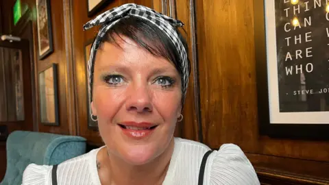 Clare is pictured looking into the camera and smiling; she is wearing a white blouse with black dungaree straps just visible on her shoulders. Her hair is held back by a black and white checked headband.

She's sat on a sofa in a bar lounge area; the walls are dark wood and panelled, and there's various pictures hanging on the walls just out of focus. 