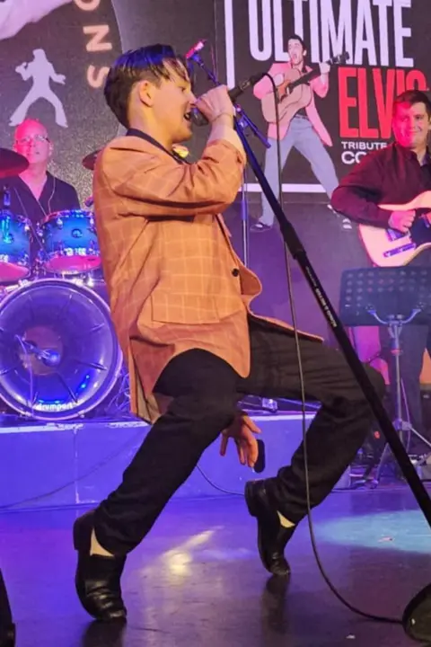 Cooper is singing on stage. He is leant back with bent knees and he is standing on his toes in a typical Elvis-style dance move.