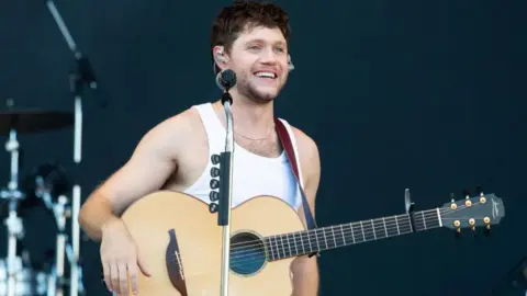 Getty Images Niall Horan performs on stage at a festival. He's a white man and is standing with a guitar on a strap around his shoulders with his right arm resting on the end of the instrument. He's wearing a white vest and is smiling looking into the distance.