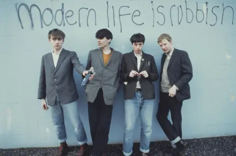 Kevin Cummins Blur's four members holding spray cans in front of graffiti on a wall reading "Modern life is rubbish"