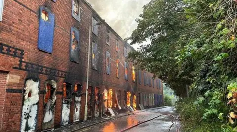 Hereford and Worcester Fire and Rescue Service A three-storey derelict red-brick building with boarded up windows. Flames can be seen through the windows of the first two storeys and fire hoses are running down the street.