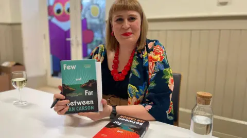 Jan Carson poses with a copy of her book Few and Far Between. She is wearing a black coat with flowers on it
