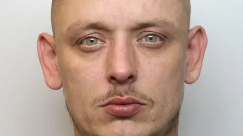A police mugshot of a man with looking at the camera, with a moustache and hazel eyes.