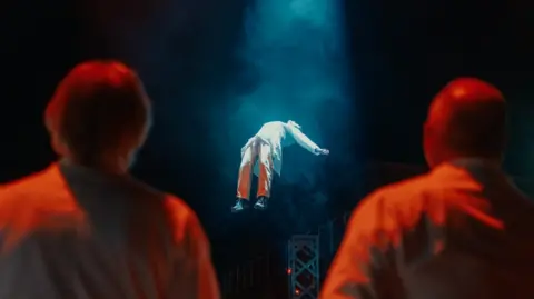 OutThere Two men with their backs to the camera watch as another man, in a white lab coat, who is floating in the air on his back with a light on him and smoke surrounding him.