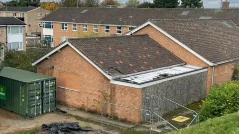 The exterior of the building with a green container next to it. It is in need of repair with missing roof tiles.