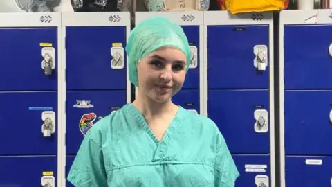 Contributed Tabatha wears surgeon scrubs and a cloth hat in a green colour. She stands in front of blue lockers.
