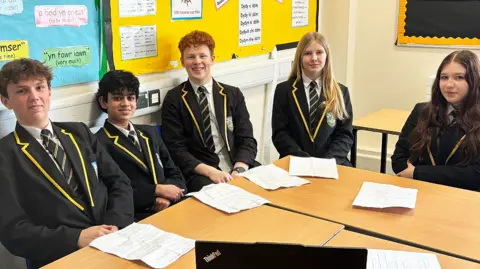 Five Year 10 students sit around two desks pushed together. They all wear a white shirt, a navy, white and gold striped tie, navy blazer with gold edging on the collar and a school badge on their left. Kit is white with short light brown hair. Jake has olive skin with short black hair. Hugo is whit with slightly curly red hair which is cropped at the sides. Cali is white with long blonde hair. Maisy is white with very long brown hair which is slightly wavy at the bottom. There are pinboards with phrases in Welsh behind them.