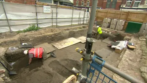 Two archaeologists work on a site in Gloucester city centre. One man is on his hands and knees, wearing a red hi vis as he digs, and another can be seen wearing a green hi vis as he stands, working the ground with a tool.