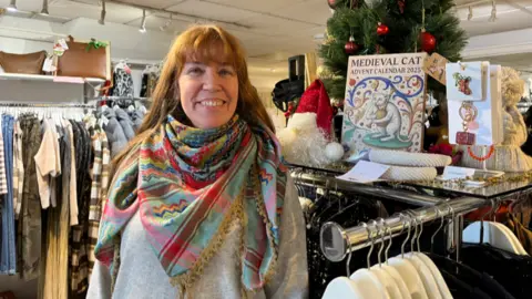 A woman inside a shop smiles at the camera. She is wearing a multi-coloured scarf and is standing next to a clothes rail. Items including keyrings and a medieval cat advent calendar are on display above the clothing on the rails. 