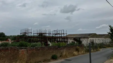 Remnants of a Victorian factory behind a brick wall. Structures pointing up to the grey sky. Buildings to the right and a road in the foreground. 