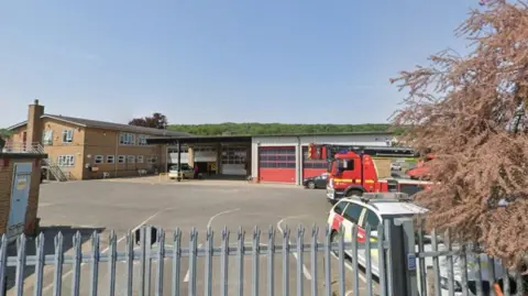 A Google Maps screengrab of the fire station. It shows a red fire engine and cars behind a grey fence. There is a large building in the background.